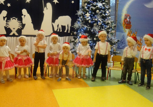 Grupa dzieci śpiewa piosenkę, w tle dekoracja świąteczna.